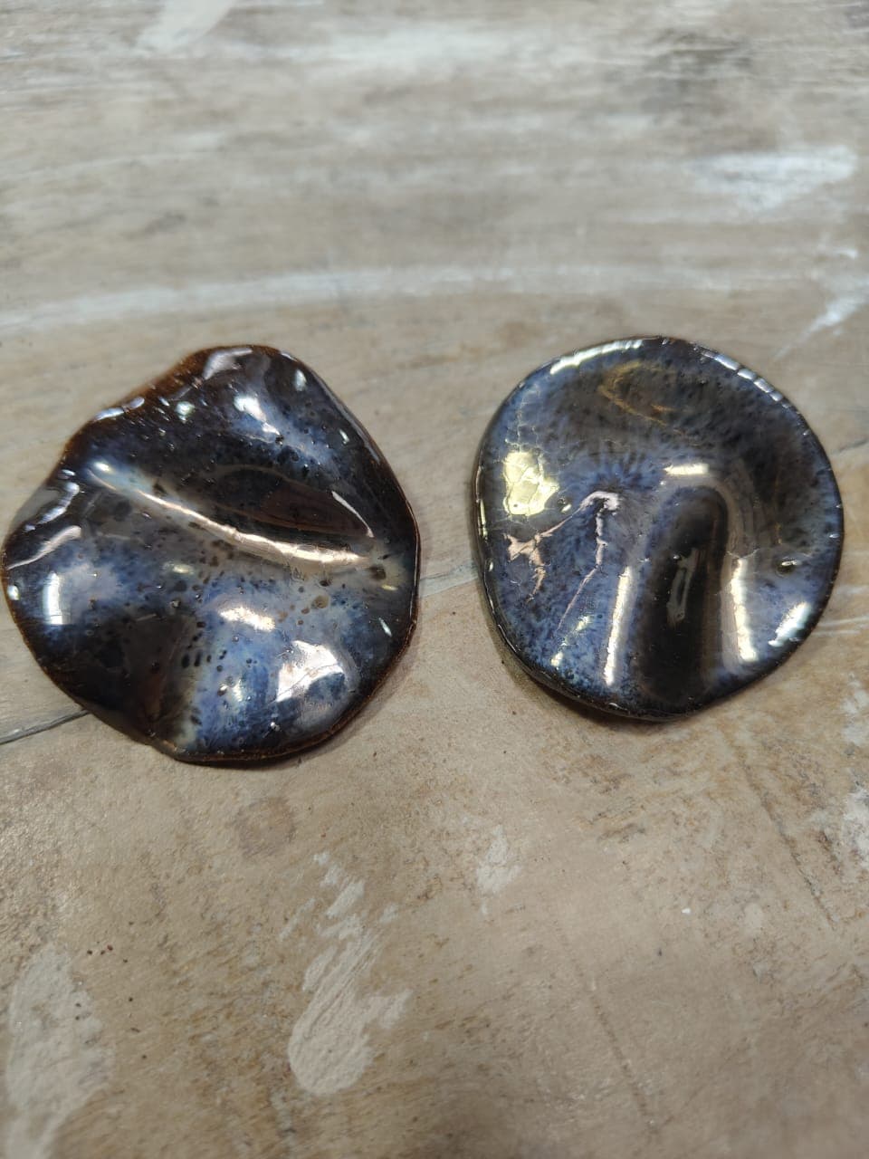 Par de brincos em disco de borda ondulada, esmalte brilhante que vai do marrom-ferrugem ao azul-violeta salpicado de preto, sobre madeira clara.